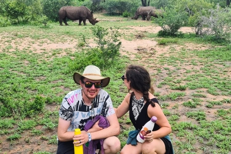 Emma and Murray crouching in the bush with two rhinos 12 feet behind them; Emma looks at the rhinos, Murray looks at the camera